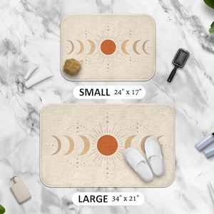 Può includere: Due tappeti da bagno beige con un design celeste con un sole e fasi lunari. Il tappeto piccolo misura 61 cm x 43 cm, e il tappeto grande misura 86 cm x 53 cm. I tappeti sono rettangolari con angoli arrotondati. Il testo "SMALL" e "LARGE" sono visibili.