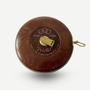 Może przedstawiać: Brązowy skórzany miernik taśmowy z mosiężną klamrą i złotą metalową pętlą. Miernik taśmowy ma nadrukowany tekst "Trade Mark" i "Made in the People's Republic of China".