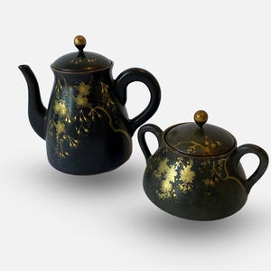 May include: A black ceramic teapot and sugar bowl set with gold floral patterns. The teapot has a curved spout and handle, and the sugar bowl has two handles. Both have lids with gold knobs.