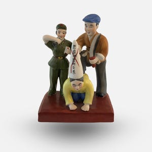 May include: A ceramic figurine depicting three figures. The central figure is kneeling and wearing a yellow shirt and a white hat with Chinese characters. The figure on the left is wearing a green military uniform and the figure on the right is wearing a brown shirt and a blue cap. The figures are standing on a red base.