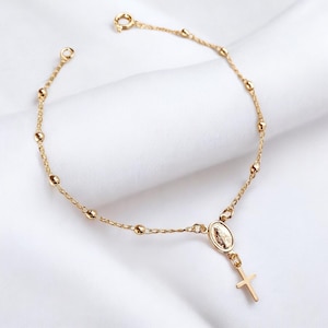 Op de afbeelding: Een gouden armband met een delicate ketting en kleine kralen. Een kleine ovale bedel met een religieus beeld hangt aan de ketting, met een kruisbedel eronder.