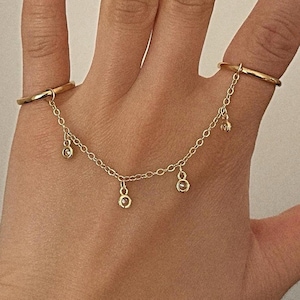 Puede incluir: Un anillo doble para dos dedos de color dorado, conectado por una delicada cadena. La cadena presenta pequeños dijes colgantes. El anillo se usa en dos dedos, mostrando su diseño único y su elegante simplicidad. La joya se presenta sobre un fondo neutro.