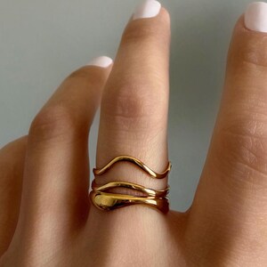 May include: Three gold rings with a fluid, organic design are stacked on a finger. The top ring has a simple wave shape, while the middle ring has a more curved form. The bottom ring has a smooth, rounded shape. The rings are a warm gold colour.