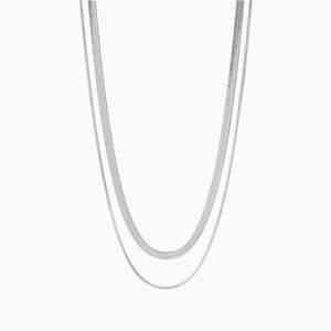 Puede incluir: Un conjunto de dos collares plateados. Los collares tienen un diseño de cadena de espiga plana. Las cadenas cuelgan verticalmente sobre un fondo blanco. Los collares son probablemente de metal.