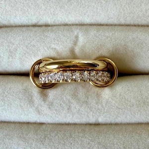 Pode incluir: Um anel de ouro com uma banda lisa e polida sobreposta a uma segunda banda cravejada com uma fila de pedras claras de corte redondo. O design inclui dois aros circulares de ouro nas laterais que unem as duas bandas numa peça única.