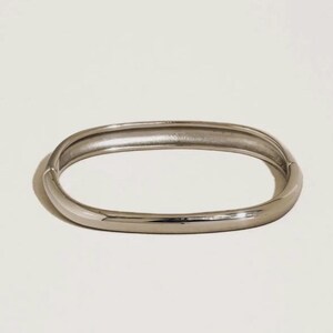 May include: A silver-colored, oval-shaped bangle bracelet. The bracelet has a smooth, polished surface and a simple, minimalist design. The bangle is a solid piece of jewelry, suitable for everyday wear.