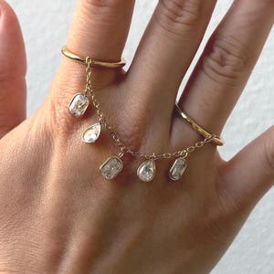 May include: A gold-colored ring set connected by a delicate chain. The chain suspends several clear, sparkling gemstones of varying shapes. The ring is worn on two fingers, showcasing the jewelry's design.