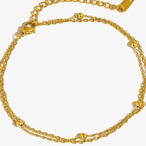 May include: A gold-toned double-strand anklet featuring delicate cable chains adorned with small, evenly spaced gold beads. The piece includes a lobster claw clasp and an adjustable extender chain with a small rectangular tag at the end.
