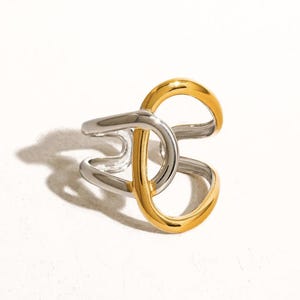May include: A two-tone ring with a modern design. The ring features a silver band that intersects with a gold-colored band, creating an open, geometric shape. The ring is set against a white background.