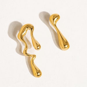 Può includere: Due orecchini astratti color oro. Un orecchino ha un design ondulato con tre sezioni arrotondate. L'altro orecchino ha una forma a goccia con la parte superiore curva. Entrambi gli orecchini sono su sfondo bianco.