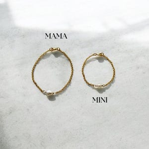 May include: Two gold-coloured chain bracelets, one larger and one smaller, each with a single white pearl. The word "MAMA" is above the larger bracelet, and "MINI" is above the smaller one. The bracelets are on a light-coloured surface.