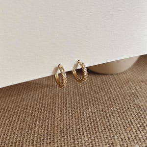 May include: A pair of small gold hoop earrings featuring a row of tiny, clear, sparkling stones embedded along the front-facing surface of each circular band. The earrings are positioned side by side, showcasing their polished metallic finish.