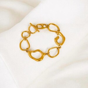 May include: Gold-toned bracelet with an abstract design. The bracelet features irregularly shaped links connected by small circular links. A toggle clasp secures the bracelet. The jewelry is displayed on a white, textured surface.