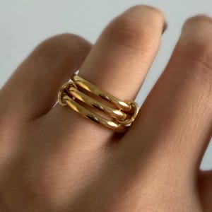 May include: A gold-colored ring with three connected bands. The ring is worn on a finger, and the bands are interlocked to create a unique design. The ring has a shiny, polished finish.