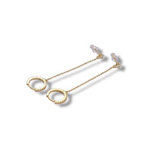 May include: A pair of gold-colored hoop earrings with a delicate chain and small clear stones. The earrings are on a white background, showcasing their design and sparkle. The earrings have a post and butterfly backing.