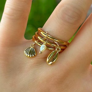 May include: Gold-toned rings stacked on a finger, each with a unique charm. Charms include a seashell, a pearl, and a cowrie shell. The rings are displayed against a blurred green background.