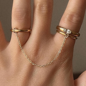 May include: A gold-toned ring set connected by a delicate chain. One ring is a simple band, while the other features a small, square-cut gemstone. The rings are worn on adjacent fingers.