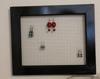 May include: A black framed wire mesh board with various pairs of earrings displayed. The earrings are made of green, red, and black beads.