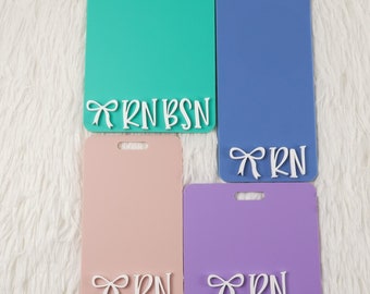 May include: Four rectangular badge holders in teal, blue, pink, and purple. Each has a white bow and the letters "RN" or "RN BSN" at the bottom. The badge holders have a hole at the top for a lanyard.