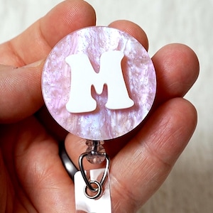 May include: A white letter "M" on a pink and white iridescent glitter background. The letter is attached to a clear plastic retractable badge holder.