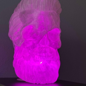 May include: A clear glass skull sculpture with a sculpted hairstyle and beard. The skull is illuminated from within with a bright pink light.