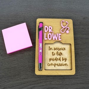 May include: A wooden desk organizer with the name "DR. LOWE" and a stethoscope design. A pink pen and a stack of pink sticky notes are also present. The organizer includes the text "In service to life, guided by compassion."