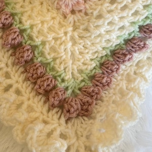 May include: A handmade crochet blanket in cream, light green, and pink. The blanket features a floral design with raised, textured details. The edges have a scalloped trim. The overall aesthetic is soft and delicate, suitable for a baby blanket or decorative throw.