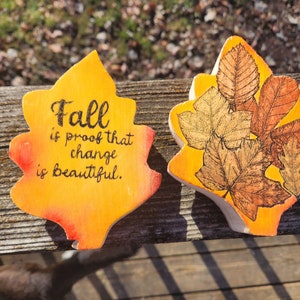 May include: Two wooden leaf-shaped signs with a fall theme. One sign is painted yellow and orange with the text "Fall is proof that change is beautiful." The other sign is painted yellow with a brown leaf design.