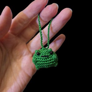 May include: A small, green crocheted frog pendant with black eyes and a stitched mouth. The frog is suspended from a green cord, creating a whimsical accessory. The background is a solid black.