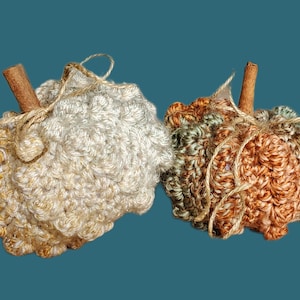 May include: Two crocheted pumpkins, one in beige and gold and the other in brown, green, and beige. Both pumpkins have a cinnamon stick stem.