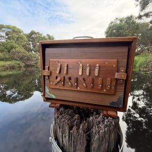 May include: A handcrafted wooden box with a hinged lid and a series of wooden toggles and brass accents. The box is a rich brown color and sits on a wooden post near a body of water. The box has a metal handle.