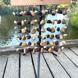 Op de afbeelding: Een houten frame met een reeks touwen die verschillende bruine en gouden zaaddozen in verschillende maten vasthouden. Het frame is gemonteerd op een zwarte metalen standaard. De zaaddozen zijn in een patroon gerangschikt en creëren een muziekinstrument.