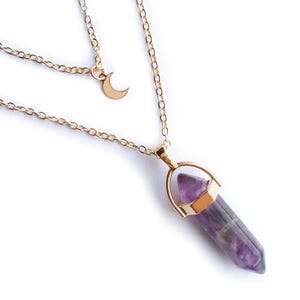 May include: A layered gold-tone necklace featuring a crescent moon charm and a purple crystal pendant. The crystal is a hexagonal shape with gold-tone metal accents. The necklace is on a white background.