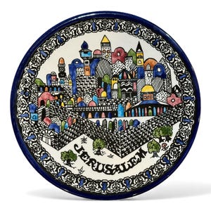 May include: A round, hand-painted ceramic plate with a dark blue rim and a detailed illustration of Jerusalem. The artwork features colorful buildings, trees, and the word "Jerusalem" in black. The plate has a decorative border.