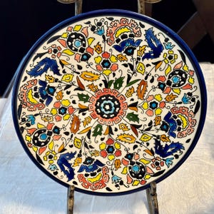 May include: A round ceramic plate featuring a vibrant, symmetrical floral pattern in shades of blue, yellow, red, green, and black against a white background. The design radiates from a central circular motif, enclosed by a solid dark blue rim.