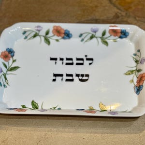 May include: White ceramic rectangular serving tray with a floral design featuring pink, blue, and green flowers. The tray has the Hebrew words "L'Chaim" and "Shabbat" written in black.