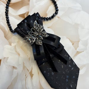 Könnte beinhalten: Eine schwarze Krawatte mit einem dezenten blauen und grauen Blumenmuster, akzentuiert durch schwarze Samtbänder und eine mittige, silberfarbene Schmetterlingsbrosche mit dunklen Kristallen. Die Krawatte ist an einer Kette aus kleinen schwarzen Perlen befestigt.