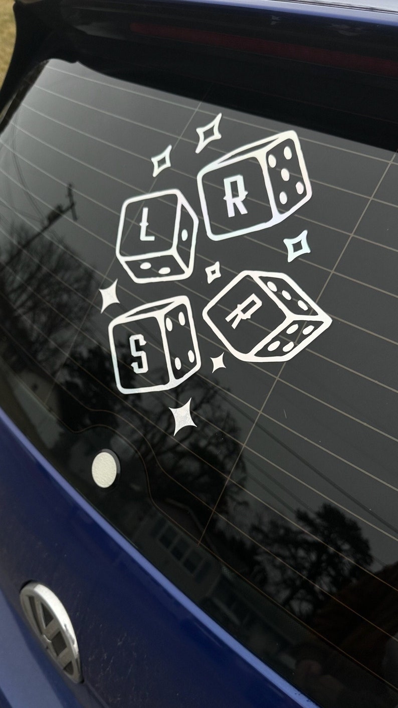 May include: A blue car with a white sticker on the back window. The sticker features four dice with the letters L, R, S, and P on them. The dice are surrounded by small stars.