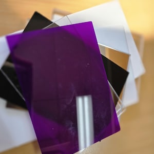May include: A stack of colorful acrylic sheets in various colors. The sheets include purple, black, white, and mirrored surfaces. The purple sheet is in the foreground, with the other sheets partially visible behind it. The sheets are on a light brown surface.