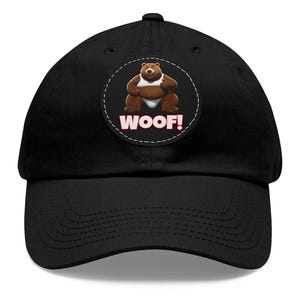 May include: Black baseball cap featuring a circular patch with a cartoon bear wearing a diaper and the word "WOOF!" in white. The bear is brown with a white diaper. The hat has a curved brim and a fabric strap closure.