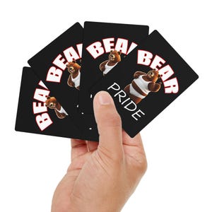 May include: A hand holding a deck of black playing cards. Each card features a cartoon bear wearing a white tank top, with the word "BEAR" in white block letters. One card reads "PRIDE".