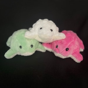 Peut inclure: Trois peluches en forme de tortues de mer. Les tortues sont couchées sur le ventre, la tête en avant. Les tortues sont blanches, vertes et roses.