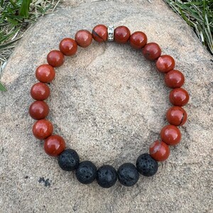 May include: A beaded bracelet featuring alternating red jasper and black lava rock beads. The red jasper beads are a deep reddish-brown color, and the lava rock beads have a porous texture. A silver-colored spacer with clear crystals adds a touch of sparkle.