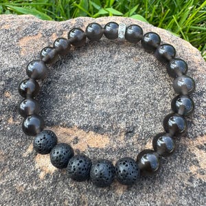 May include: A dark gray beaded bracelet featuring a mix of smooth, polished beads and textured, porous black beads. A small silver-colored spacer is also included in the design. The bracelet is displayed on a gray stone surface with green grass in the background.