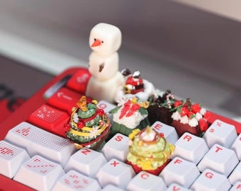 Könnte beinhalten: Eine rot-weiße mechanische Tastatur mit weißen Tastenkappen und roter Beschriftung. Miniatur-Weihnachtsdekorationen, darunter ein Schneemann und dekorierte Bäume, sind auf der Tastatur platziert und schaffen eine festliche Urlaubsszene.