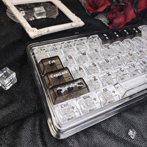 May include: A transparent mechanical keyboard with clear keycaps and black accent keys. The keyboard has a clear frame and several dark-coloured keycaps with white text and a barbed wire design. The keyboard is on a black surface.