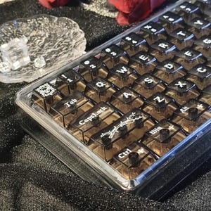 Puede incluir: Un conjunto de teclas de teclado negras translúcidas con letras y símbolos blancos. Las teclas están dispuestas en un contenedor de plástico transparente. Algunas teclas tienen diseños decorativos de calaveras y espinas.