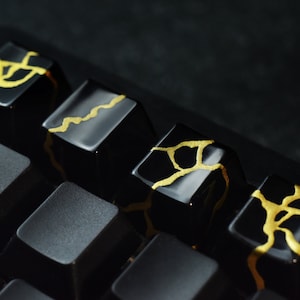 Peut inclure: Quatre touches de clavier noires avec des accents dorés. Les touches ont un design craquelé ou marbré avec des lignes et des tourbillons dorés. Les touches sont disposées en rangée sur un clavier noir.