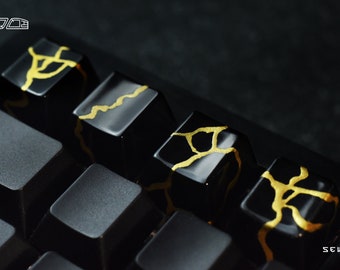 May include: Four black keyboard keycaps with gold accents. The keycaps have a cracked or marbled design with gold lines and swirls. The keycaps are arranged in a row on a black keyboard.