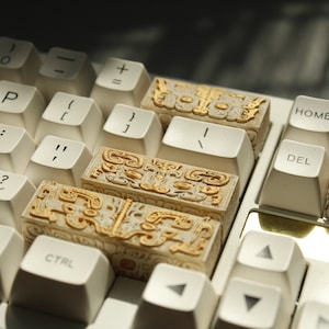 May include: A white keyboard with gold keycaps featuring intricate designs. The keycaps are in the shape of small, rectangular blocks with raised, detailed patterns. The keyboard has standard keys, including 'Home', 'Page Up', 'Del', 'Menu', and 'Ctrl'.